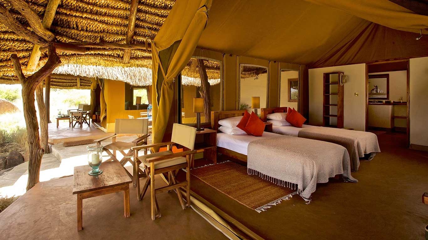 Twin tent under a thatched roof at Elewana Lewa Safari Camp, with two beds, a desk and a veranda with a daybed