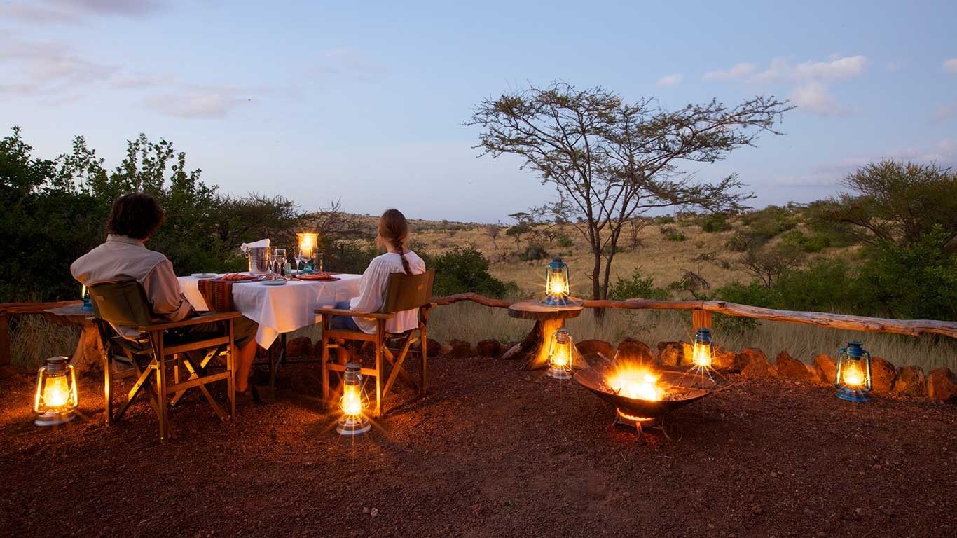 Couple dining by lantern light beside a fire pit at Elewana Lewa Safari Camp, with acacias against the evening sky