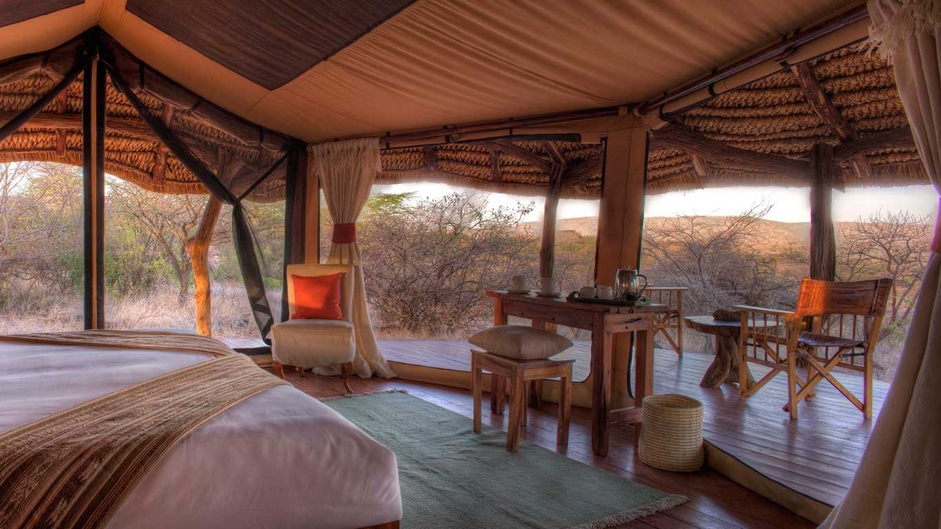 Tent interior at Elewana Lewa Safari Camp with a bed, armchair and desk, its mesh walls open to dry acacia bush