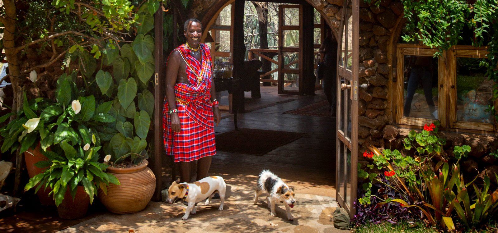 A Maasai host in a red shuka at the doorway of the main house at Lewa Wilderness, among potted plants, with two small dogs