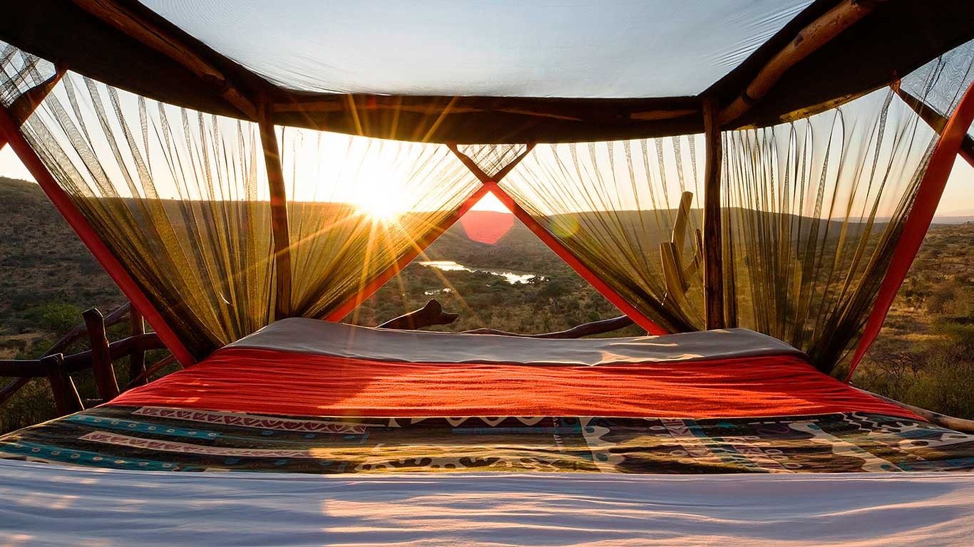 Sun rising beyond the netting of an open-air bed with a red blanket at Loisaba Star Beds