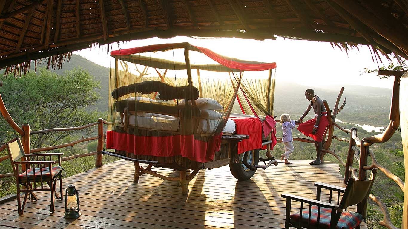 Wheeled star bed with a red blanket and netting on a wooden deck under thatch at Loisaba Star Beds