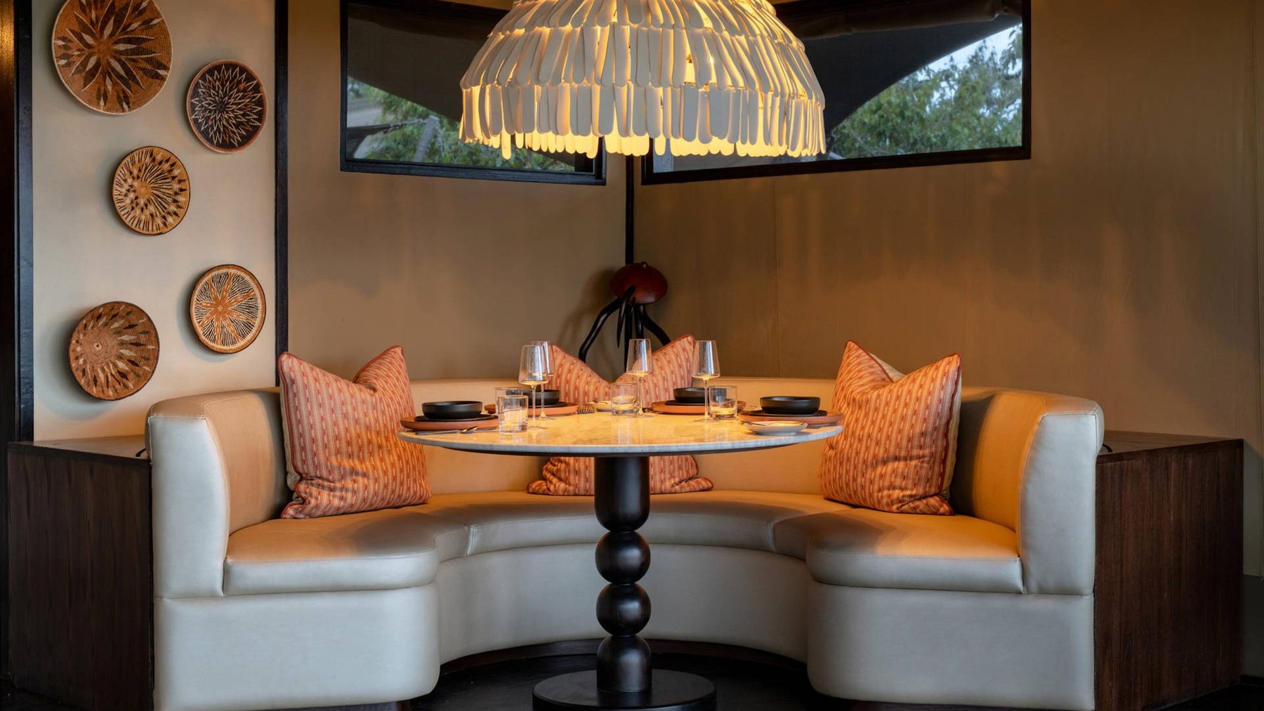Curved booth with orange cushions around a table set for dinner under a woven pendant light at Mahali Mzuri