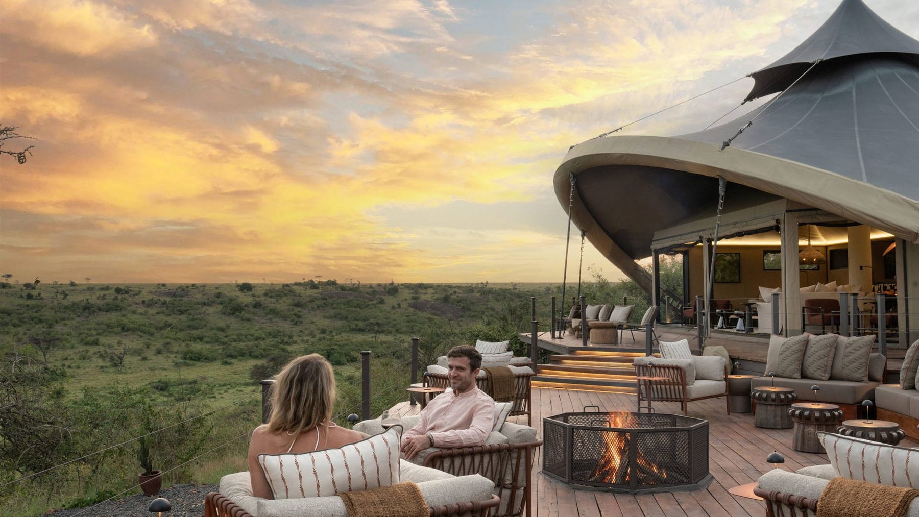 Two guests relaxing by a fire pit on the deck at Mahali Mzuri as the sun sets over the conservancy