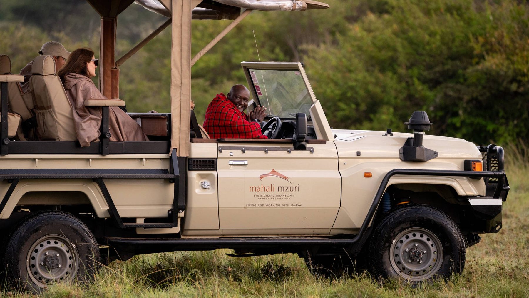 Guide at the wheel of an open Mahali Mzuri game drive vehicle, with a guest seated in the back