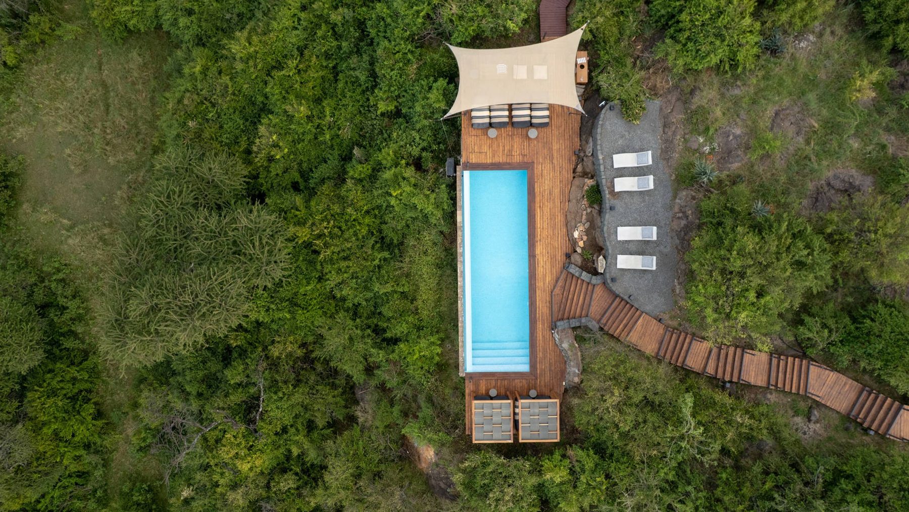 Aerial view of Mahali Mzuri’s long pool and loungers, linked by a wooden walkway through thick green bush