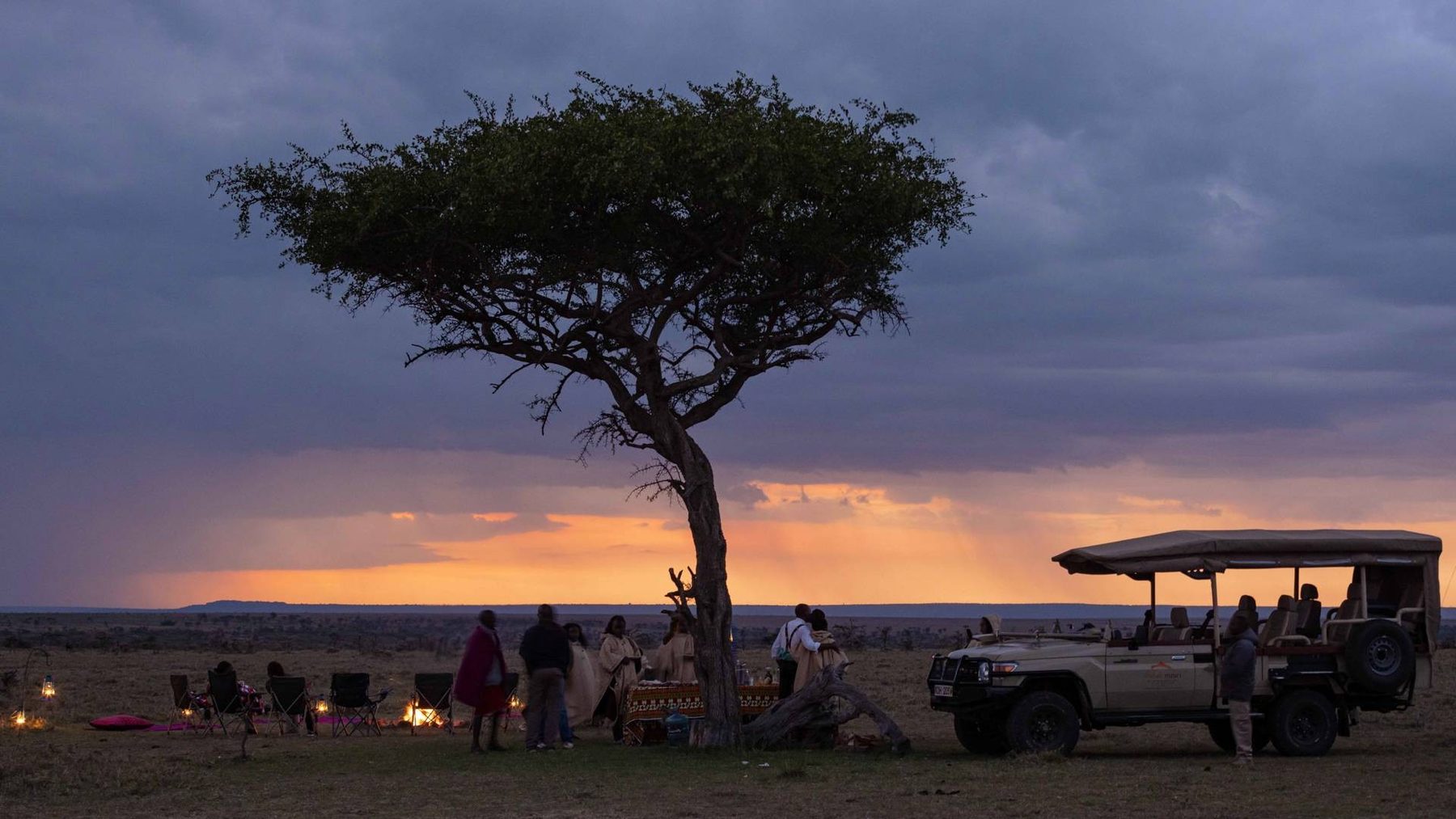 Guests gathered under a lone acacia beside a game drive vehicle as a storm sky glows orange at sunset