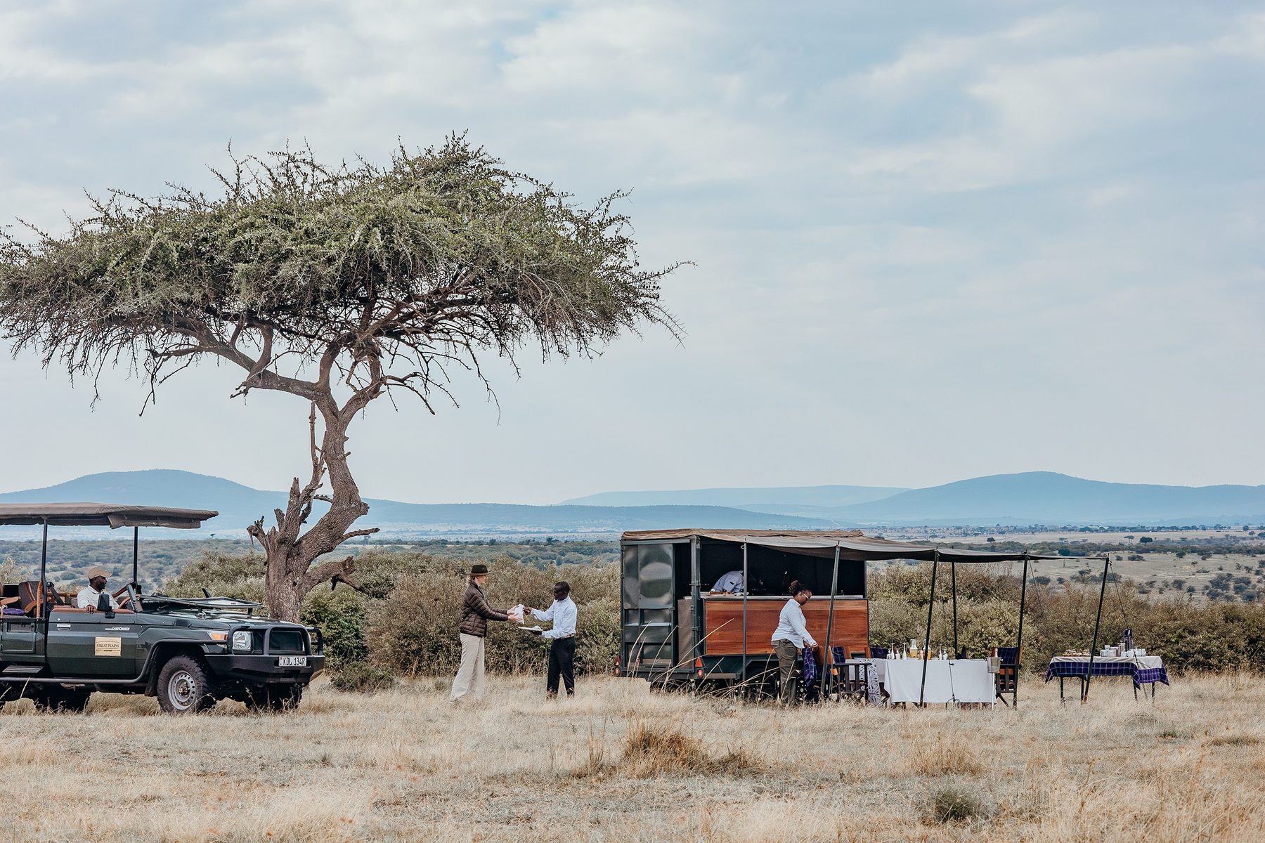 Bush breakfast laid under an acacia tree on the plains, with a game drive vehicle and kitchen trailer beside it