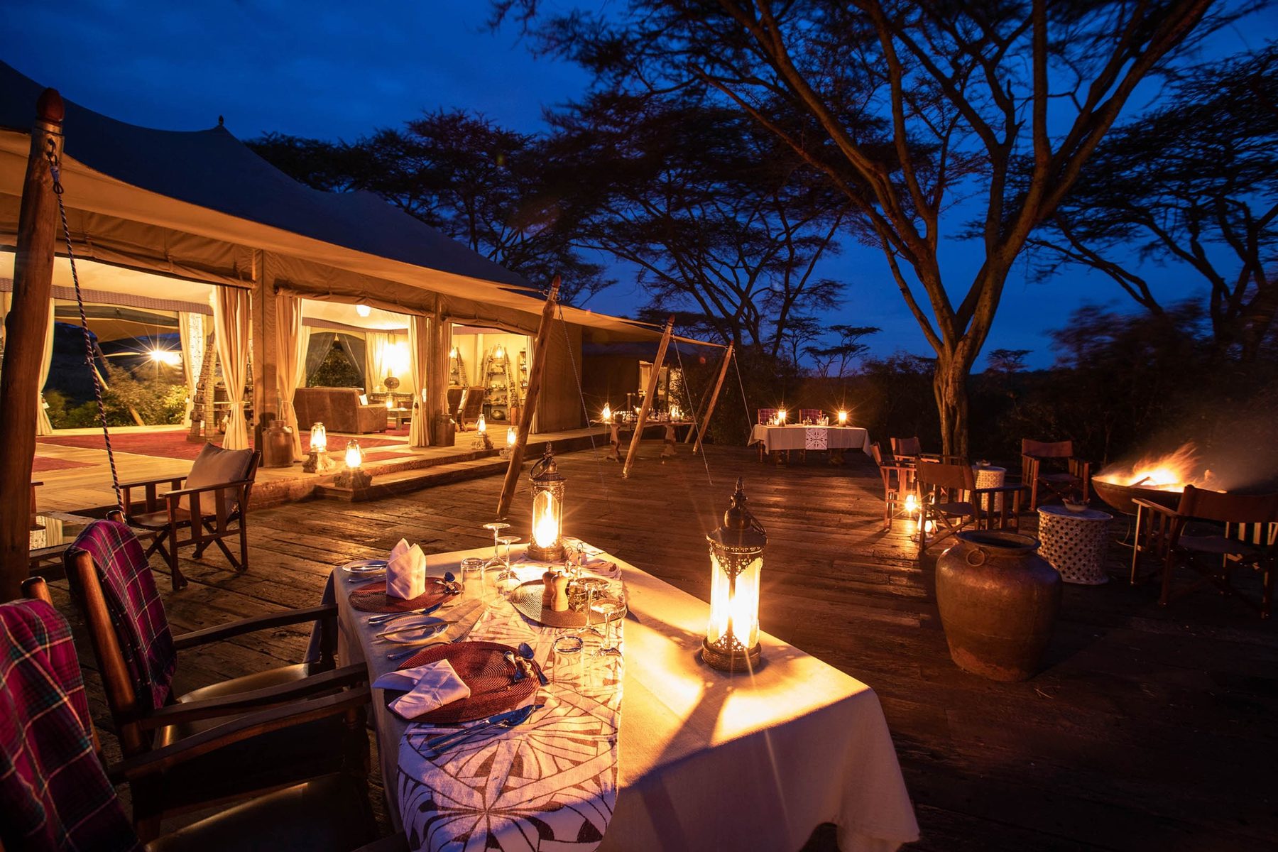 Candlelit dinner table on the deck at Mara Nyika Camp at night, with lit tents beneath acacia trees