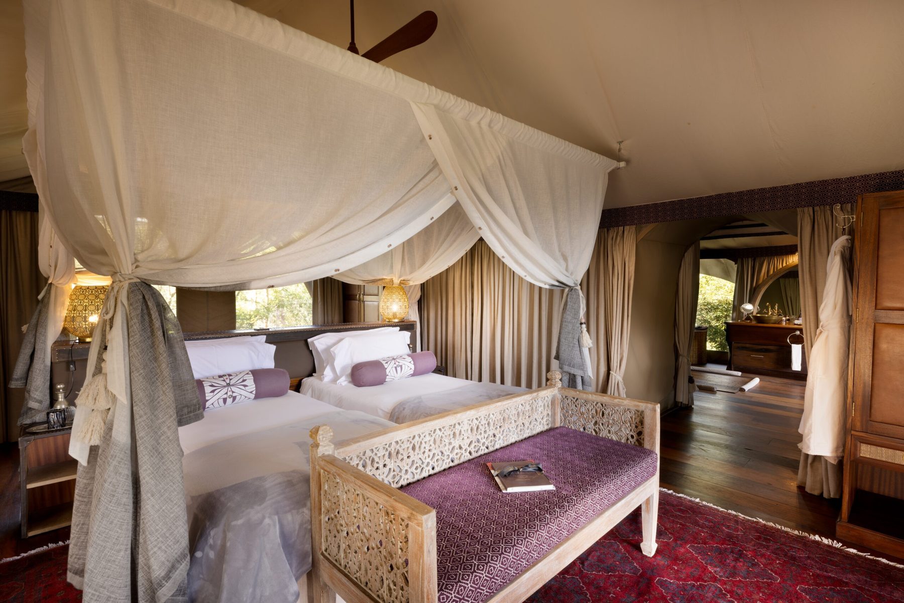 Tent at Mara Nyika Camp with a canopied bed, carved bench and Persian rug, opening onto a dressing area