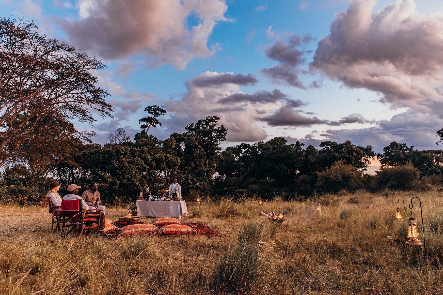 Guests at a table set for a bush meal in tall grass near Mara Plains Camp, with lanterns and rugs laid out