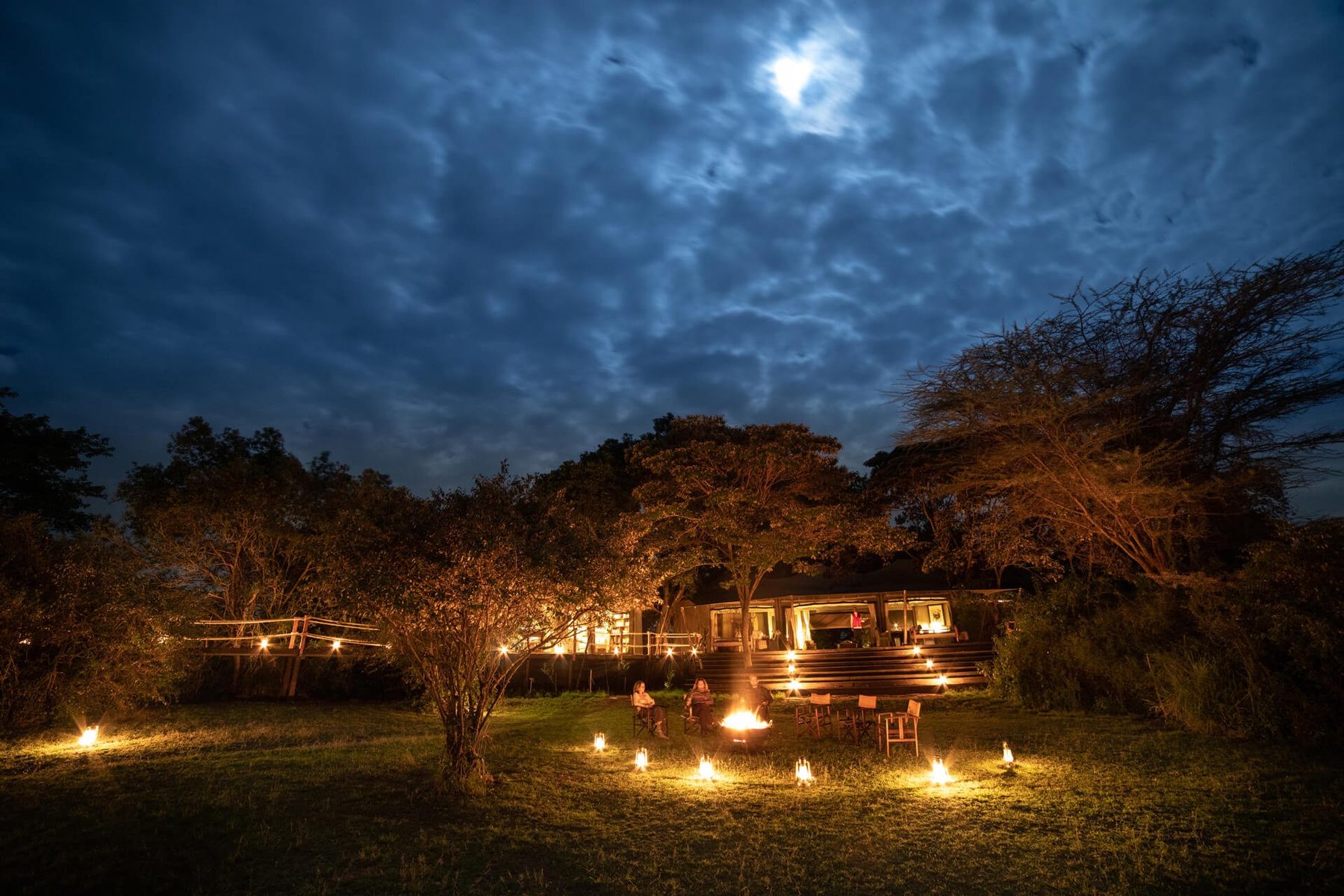 Mara Plains Camp at night, with lanterns glowing beneath the trees under a moonlit, cloudy sky