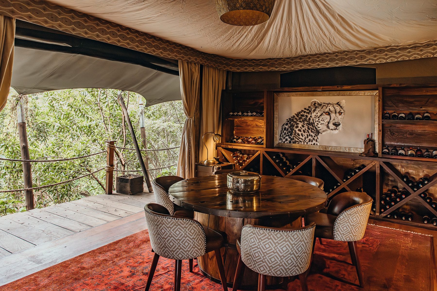 Dining tent at Mara Plains Camp with a round table, a wine rack and a framed cheetah print, opening onto trees