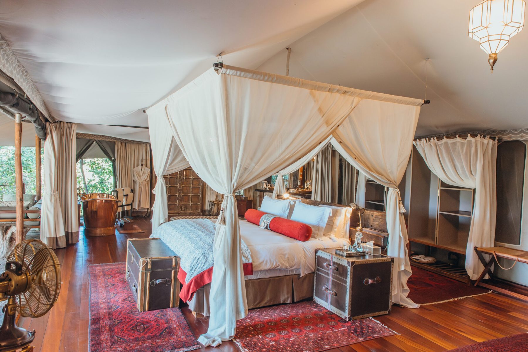 Canvas suite at Mara Plains Camp with a netted four-poster bed, travel-trunk bedside tables and red rugs on wooden floors