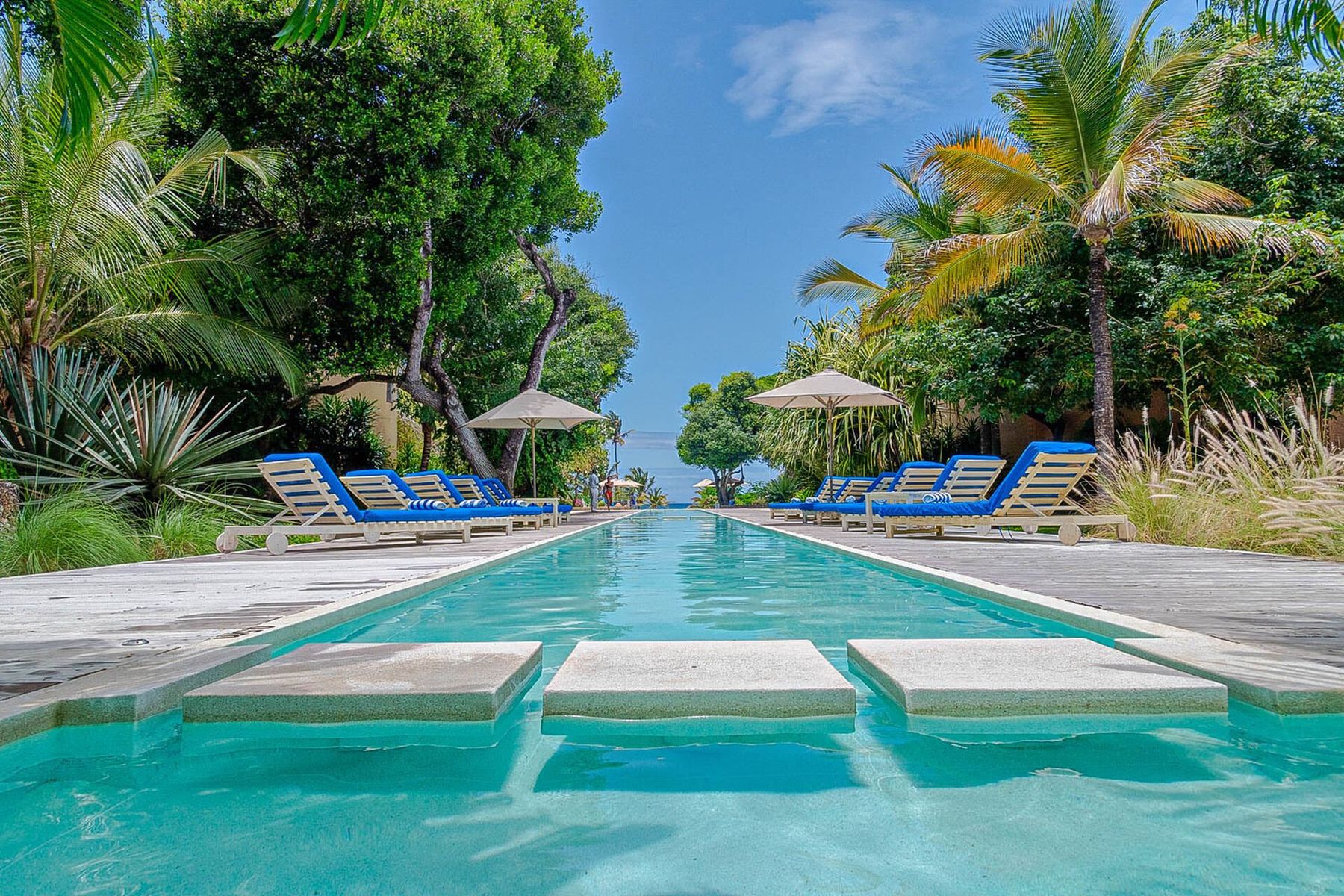 A long pool with stepping stones between rows of blue loungers and palms, leading towards the sea at Medina Palms