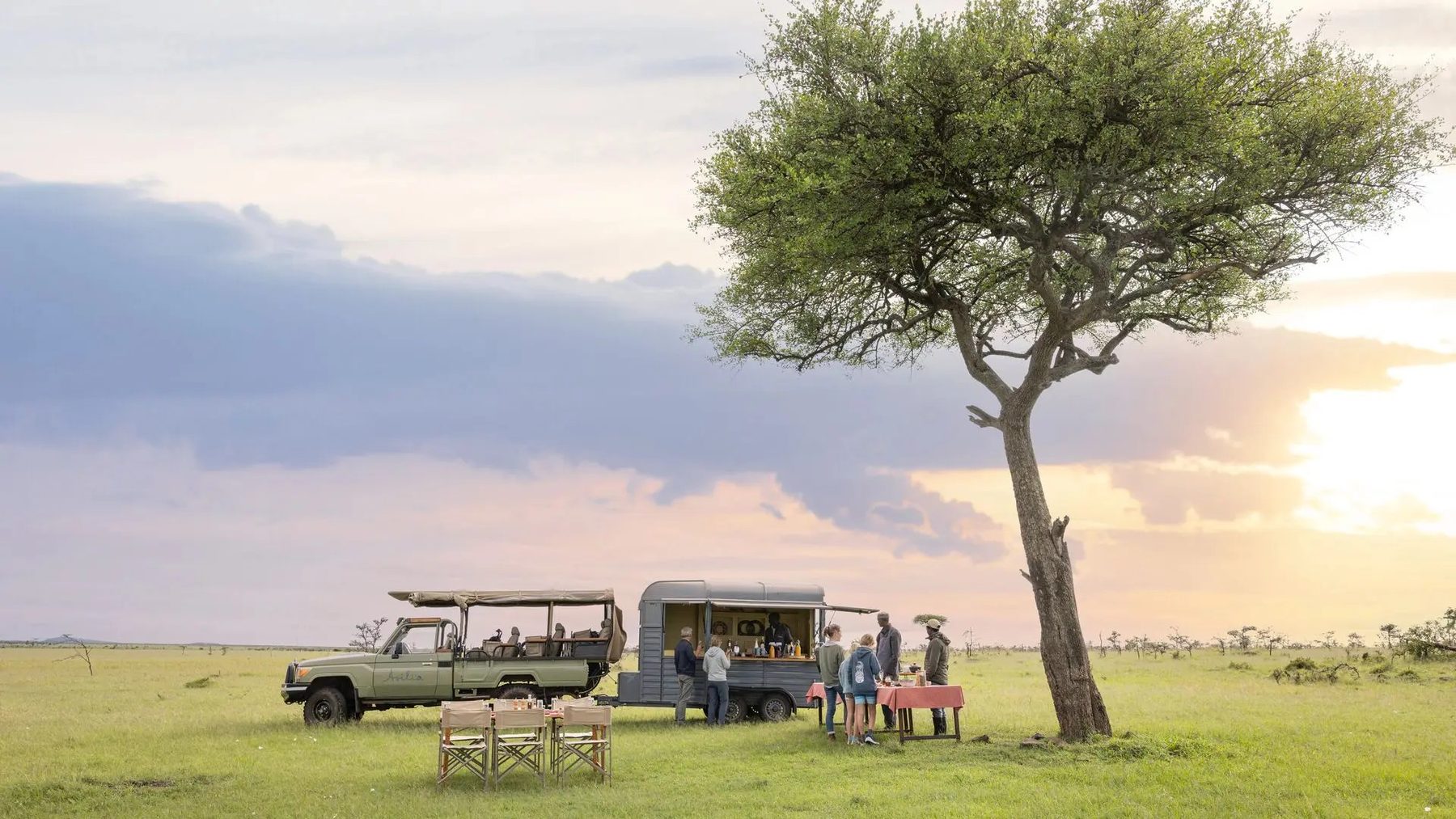 Safari vehicles and a breakfast table set beside a lone acacia on open grassland near Naboisho Camp at sunrise