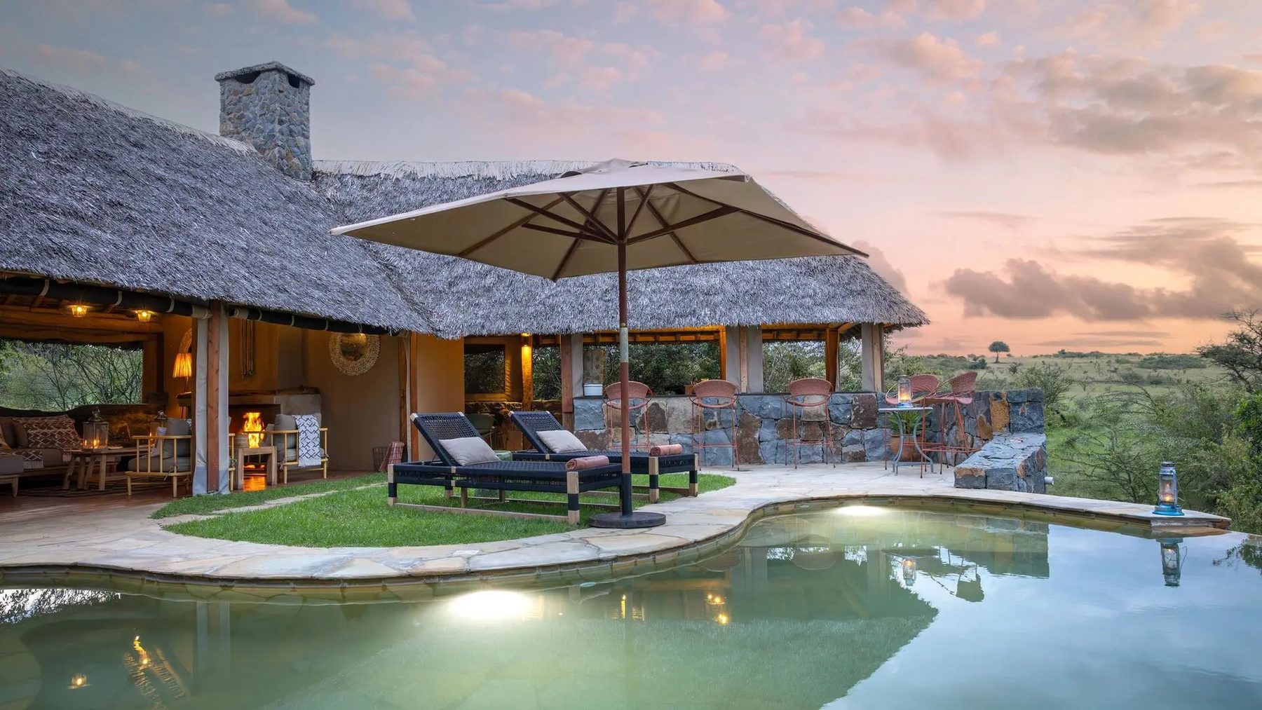 Swimming pool with loungers and a large umbrella in front of the thatched main building at Naboisho Camp at dusk