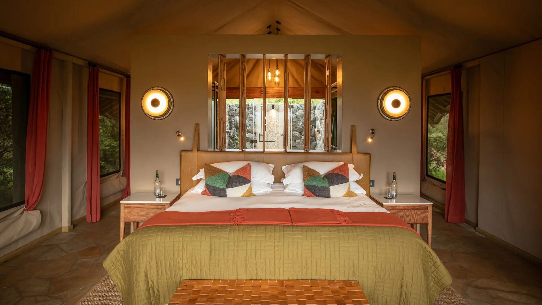 Tent at Naboisho Camp with a large bed under patterned cushions, round wall lamps and red curtains on either side