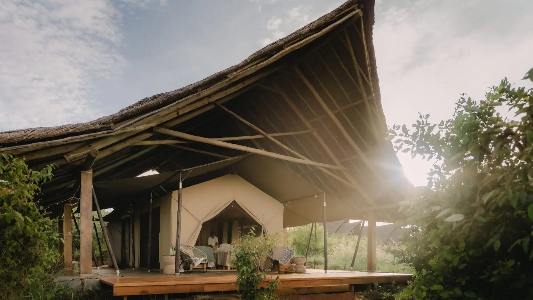 Canvas tent with a sweeping thatched roof and wooden deck among trees at Naboisho Camp, lit by low sun