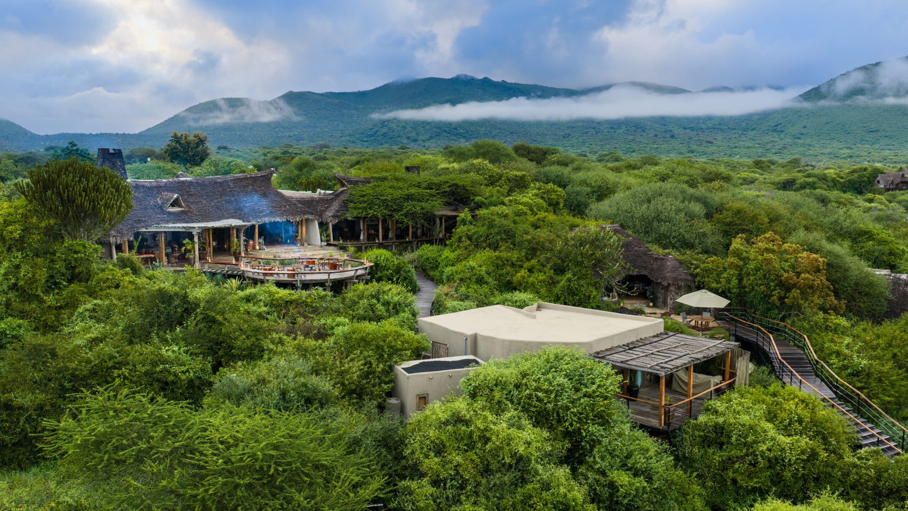 Aerial view of the thatched buildings of ol Donyo Lodge in green bush, with the Chyulu Hills behind