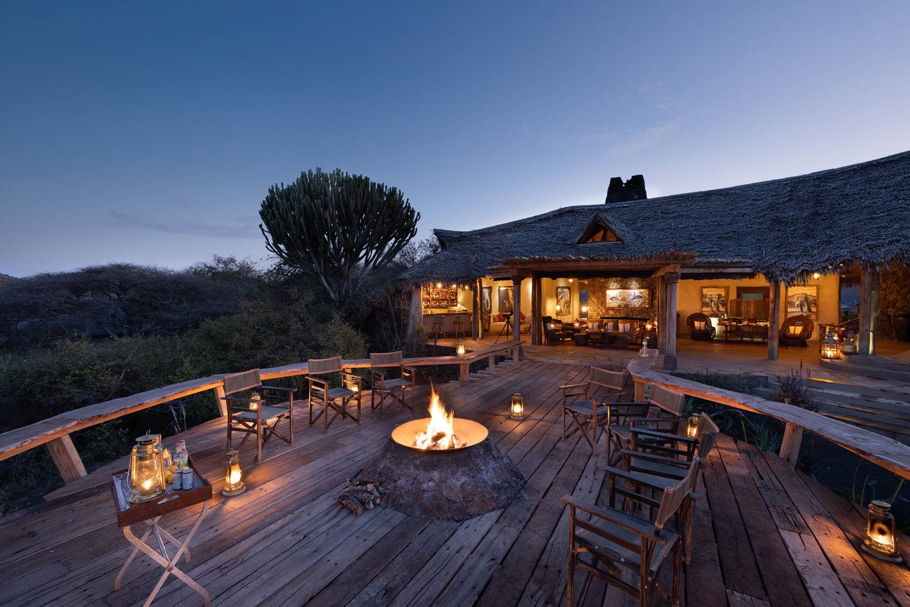 Fire pit and chairs on a wooden deck outside the lit thatched lodge at ol Donyo Lodge at dusk