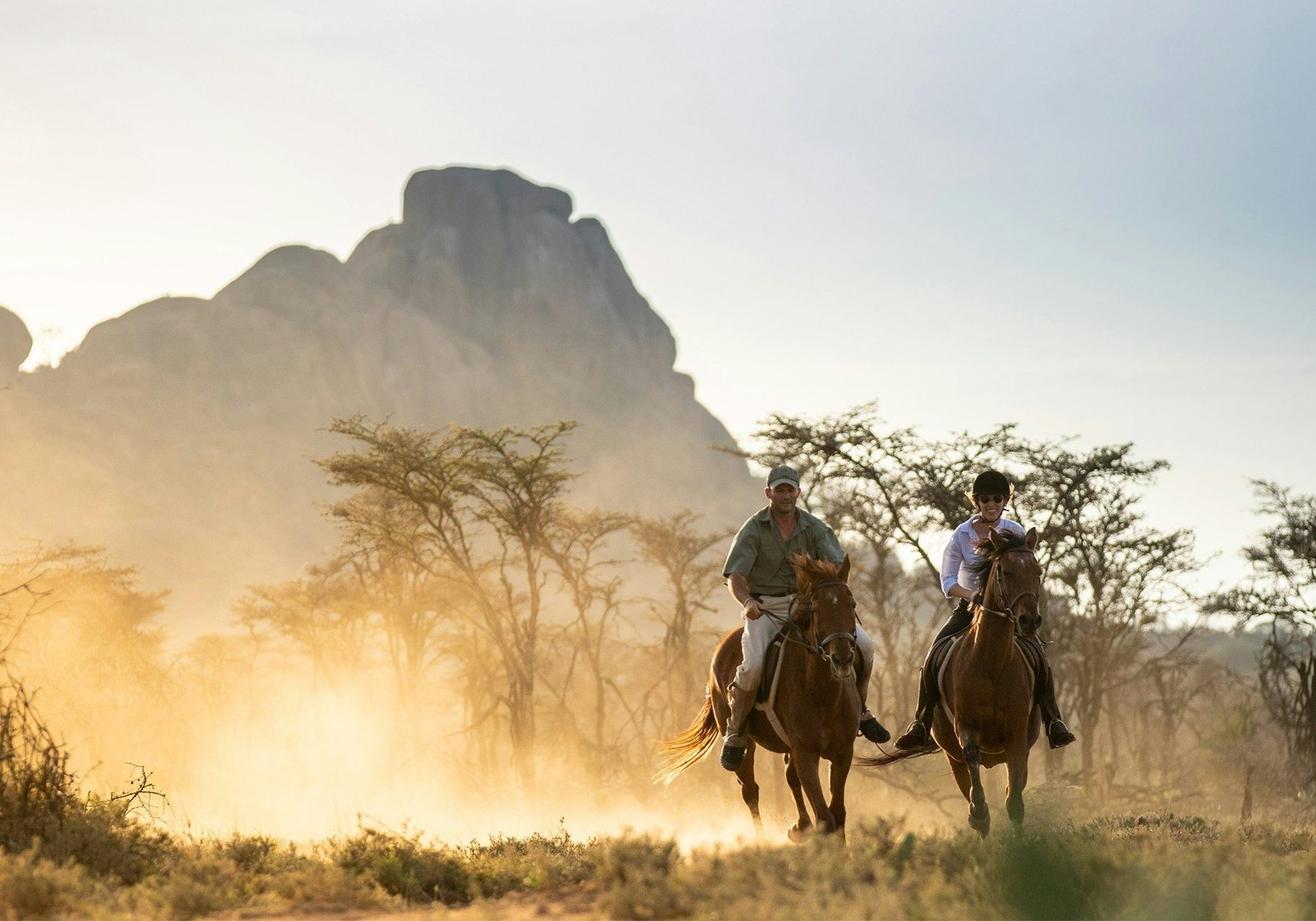 Two riders on horseback kicking up golden dust among acacias, with a rocky peak behind at Ol Jogi