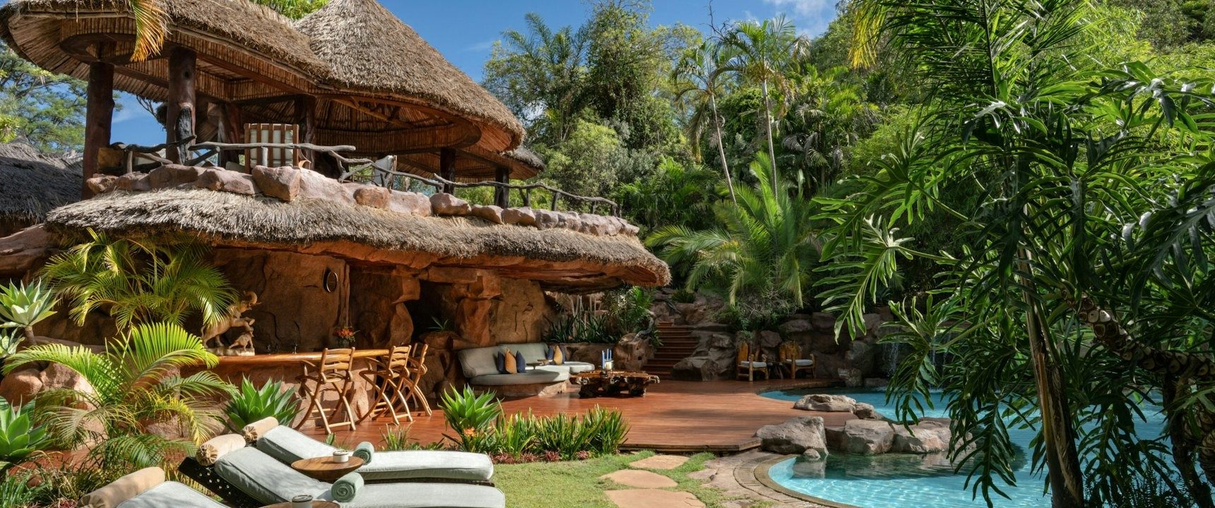 Pool with rock edges and loungers among palms and thatched buildings in the gardens at Ol Jogi