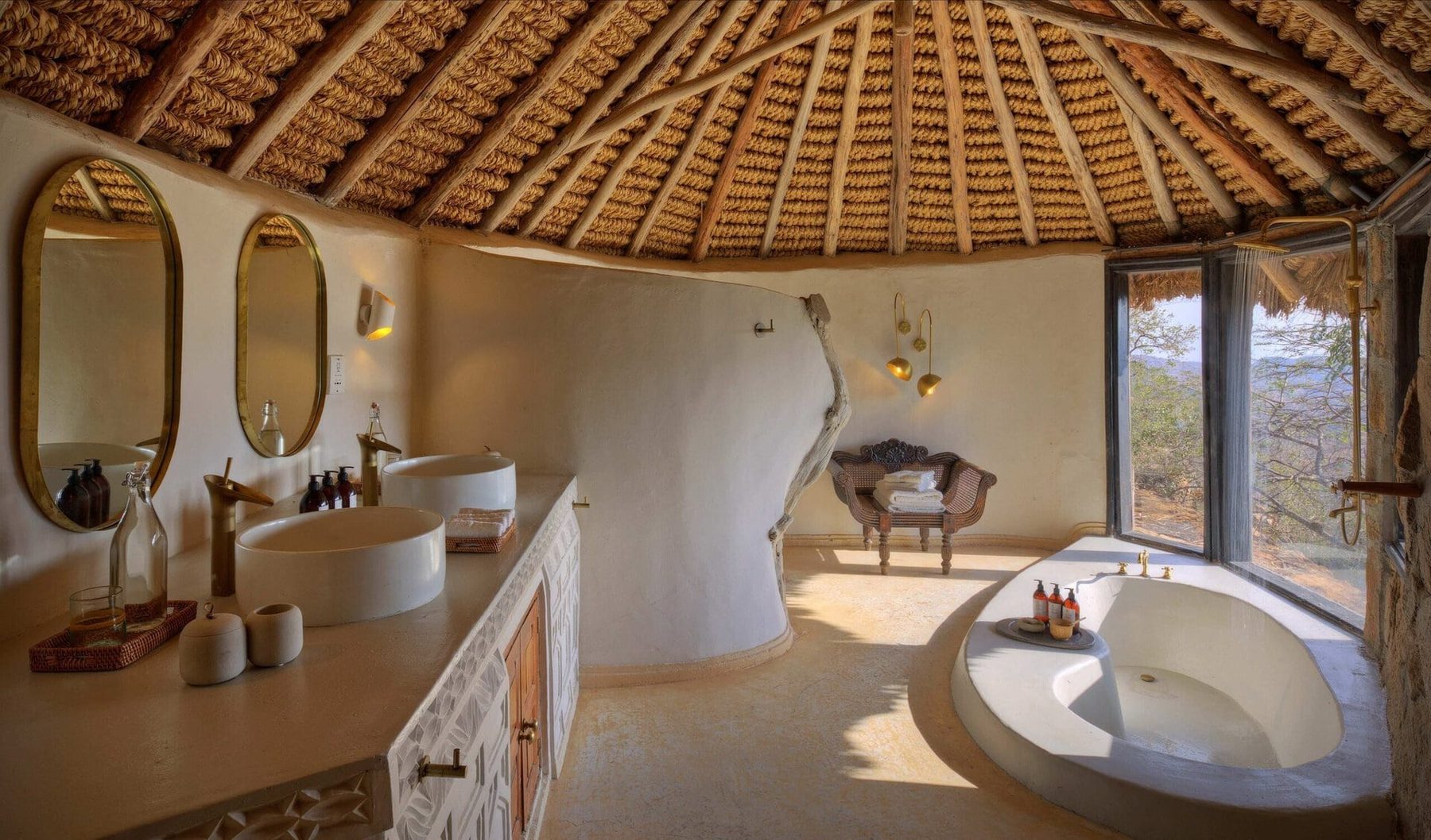 Thatched bathroom at Ol Lentille with oval mirrors over twin basins and a round tub beside the windows