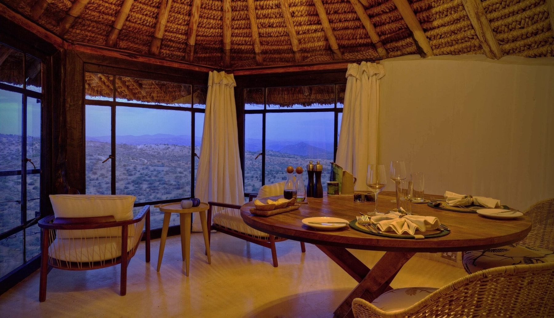 Round dining table laid for dinner in a thatched room at Ol Lentille, with windows open onto hills at dusk