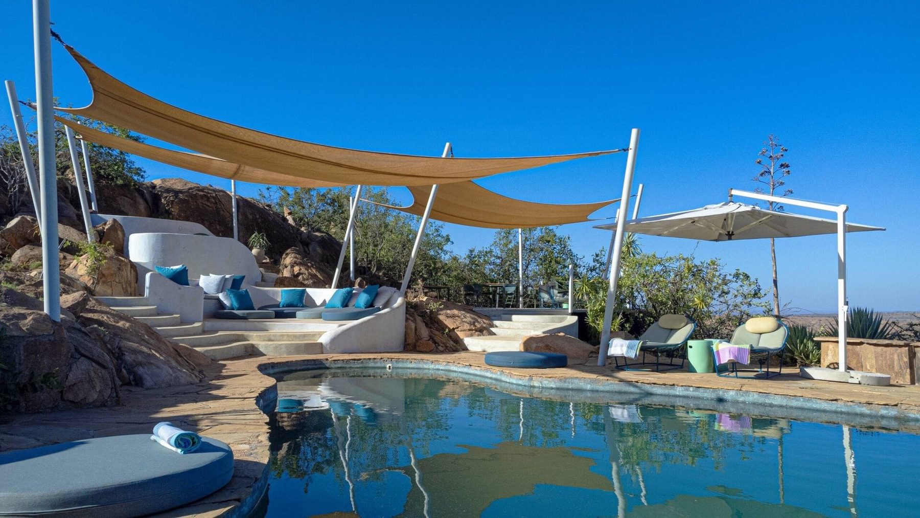 Swimming pool at Ol Lentille beneath white shade sails, with loungers on the rocky terrace above