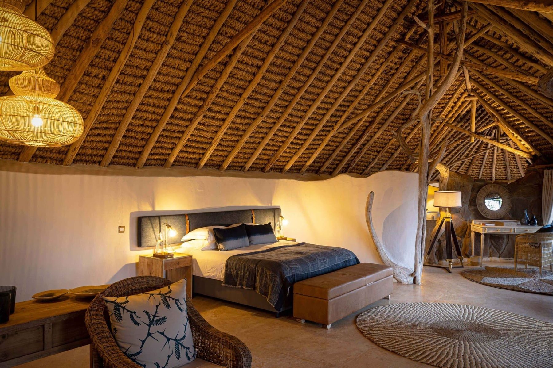 Villa bedroom at Ol Lentille under a high thatched roof, with a large bed, armchairs and a desk lit by lamps