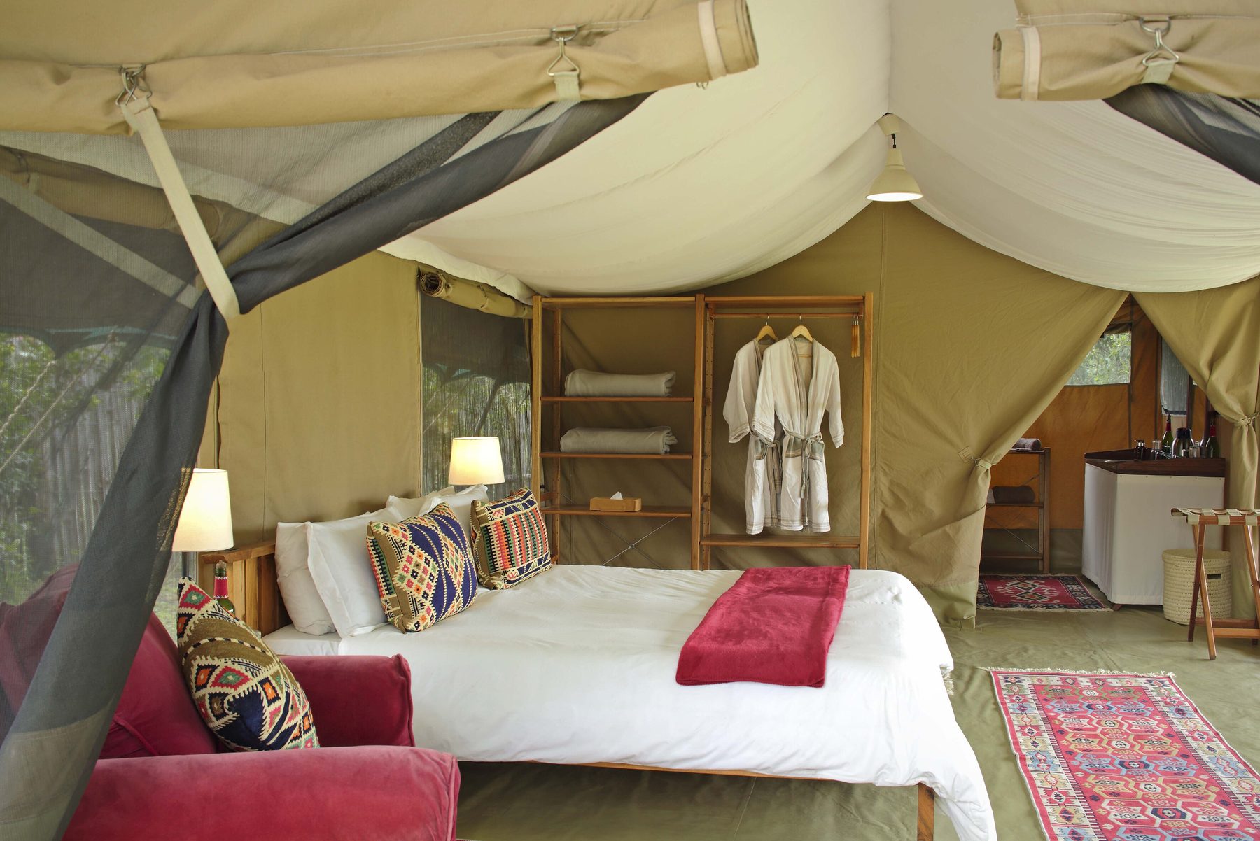 Tent at Ol Pejeta Bush Camp with a double bed, patterned cushions, a red throw and bathrobes hanging on open shelves