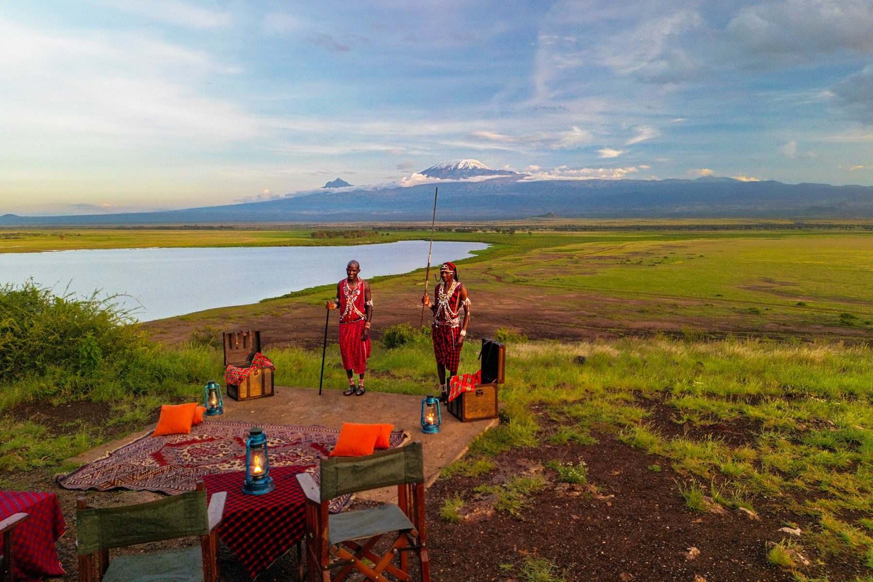 Table set with lanterns and red cloths beside a lake, with two Maasai hosts and distant Kilimanjaro, near Ol Tukai Lodge