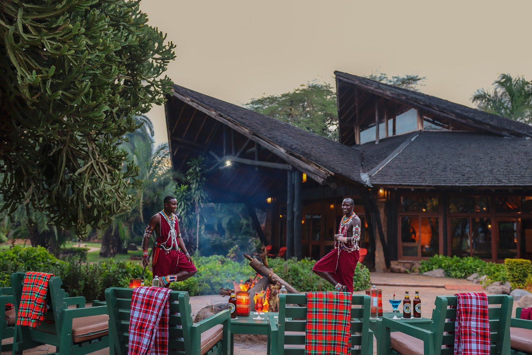 Maasai men tending a fire beside a terrace of green chairs and red blankets in front of Ol Tukai Lodge