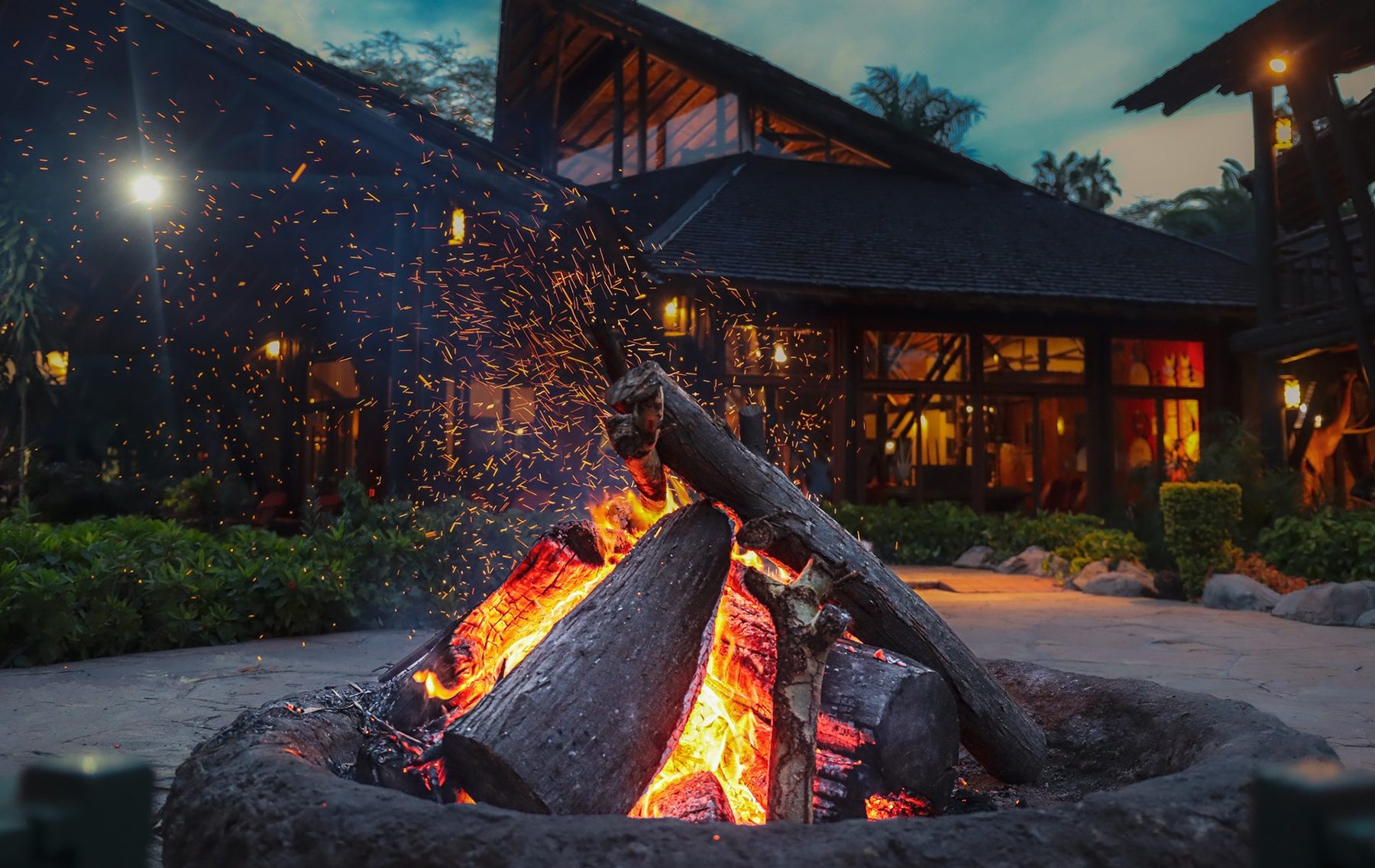 Log fire blazing in a stone fire pit in front of the lit main building of Ol Tukai Lodge at dusk