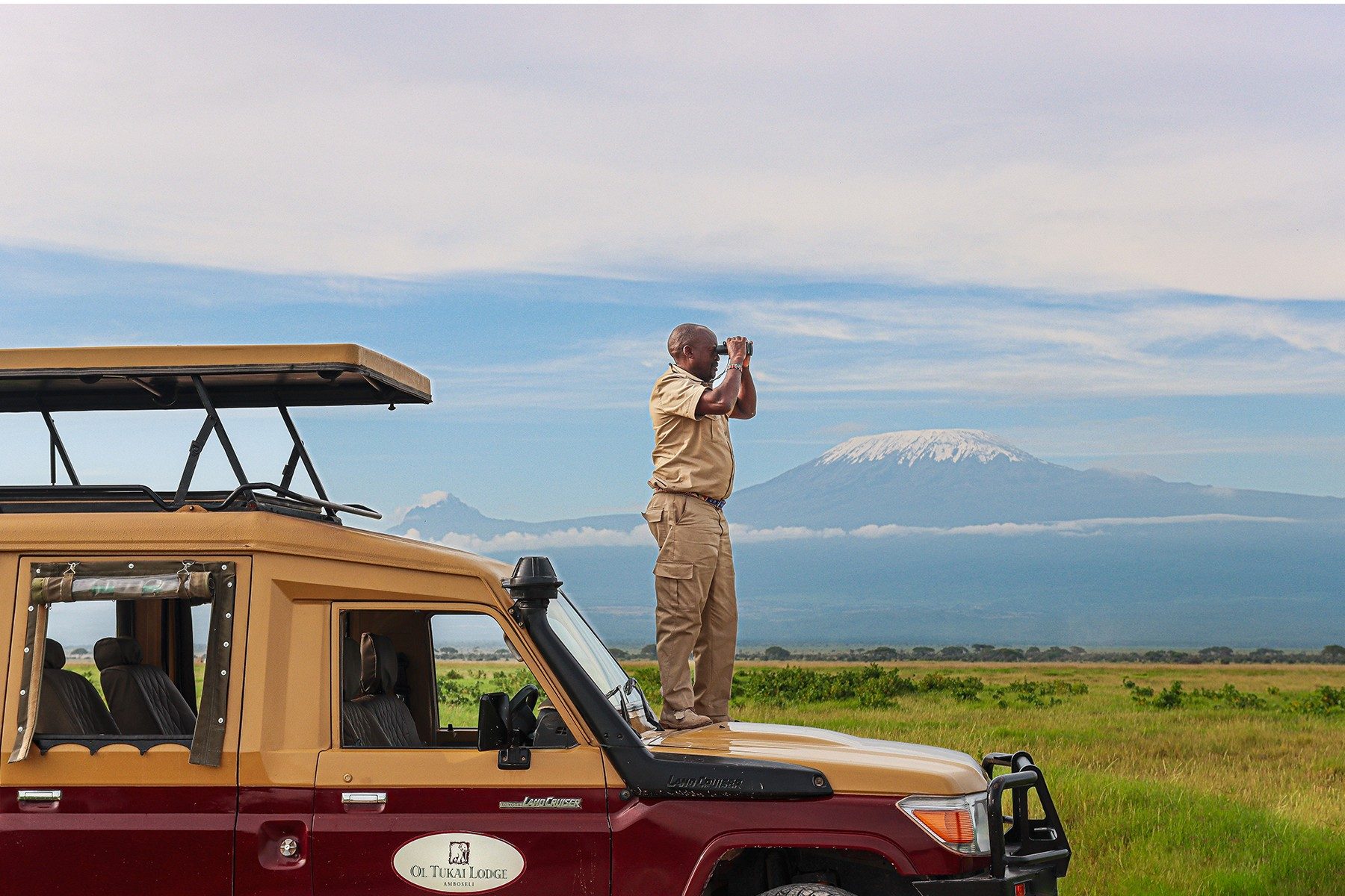 Guide standing on the bonnet of a safari vehicle with binoculars, Kilimanjaro on the horizon, near Ol Tukai Lodge