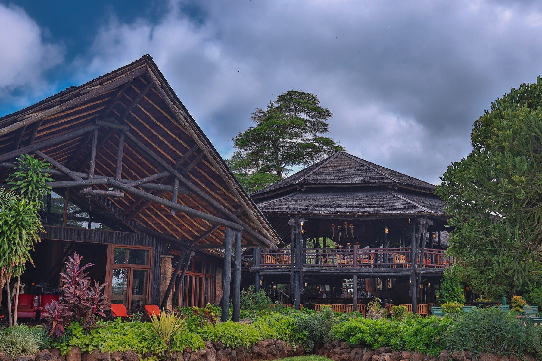 Main building of Ol Tukai Lodge with steep timber roofs, wooden balconies and gardens under a cloudy sky