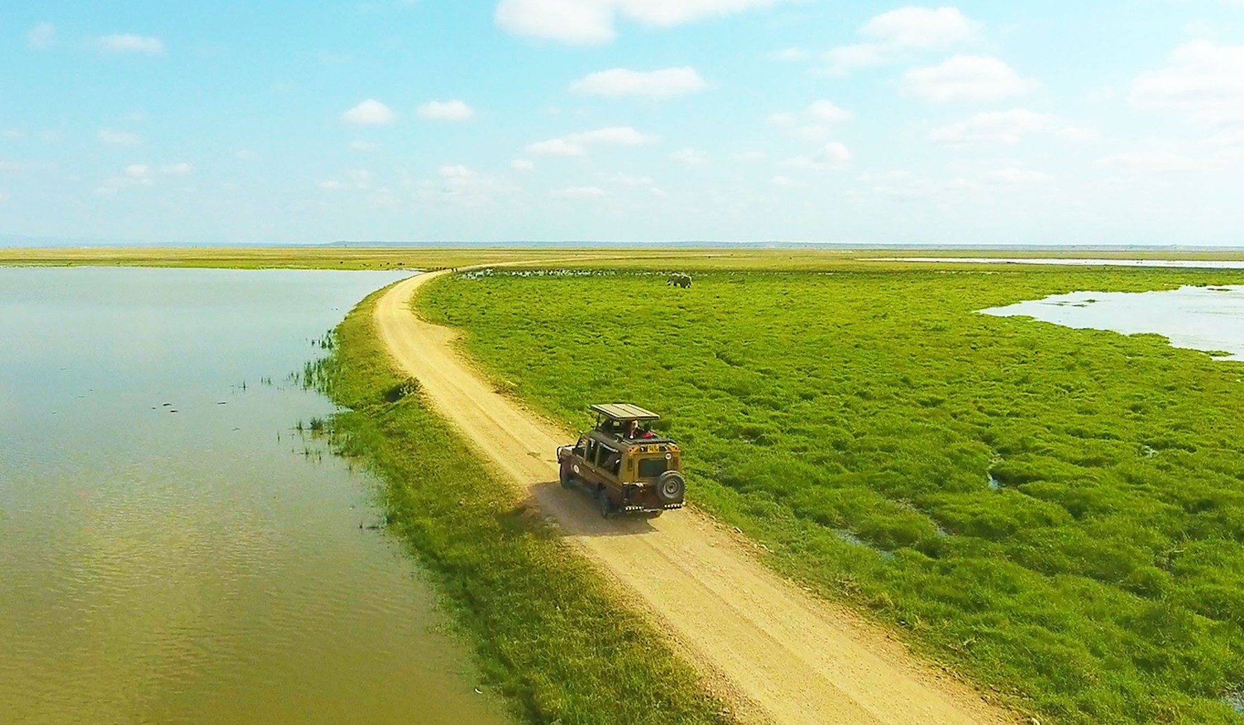 Aerial view of a safari vehicle on a raised track between still water and green marsh near Ol Tukai Lodge