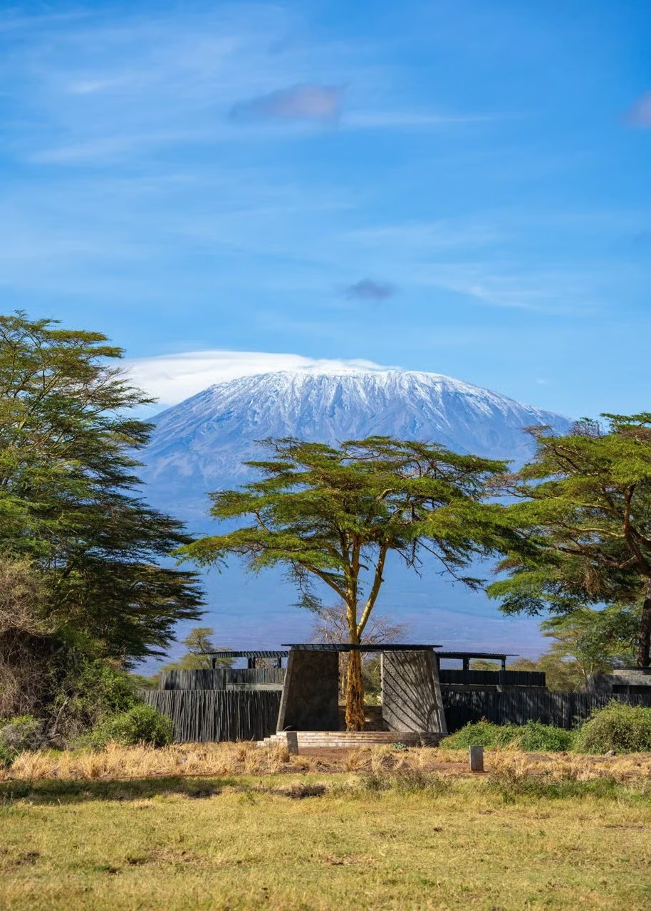 Snow-capped Kilimanjaro rising behind fever trees and a stone entrance at Angama Amboseli