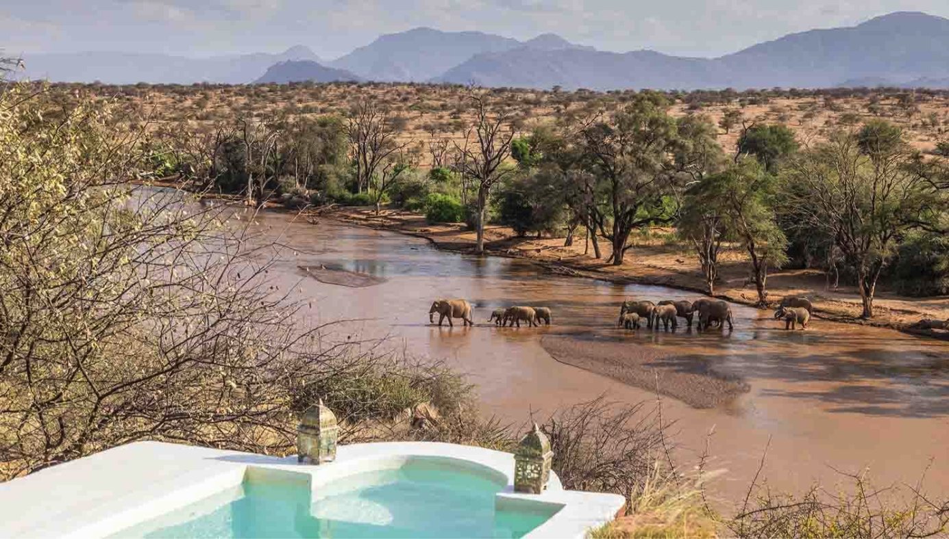 Elephants crossing a wide river below a plunge pool, with mountains behind, at Sasaab