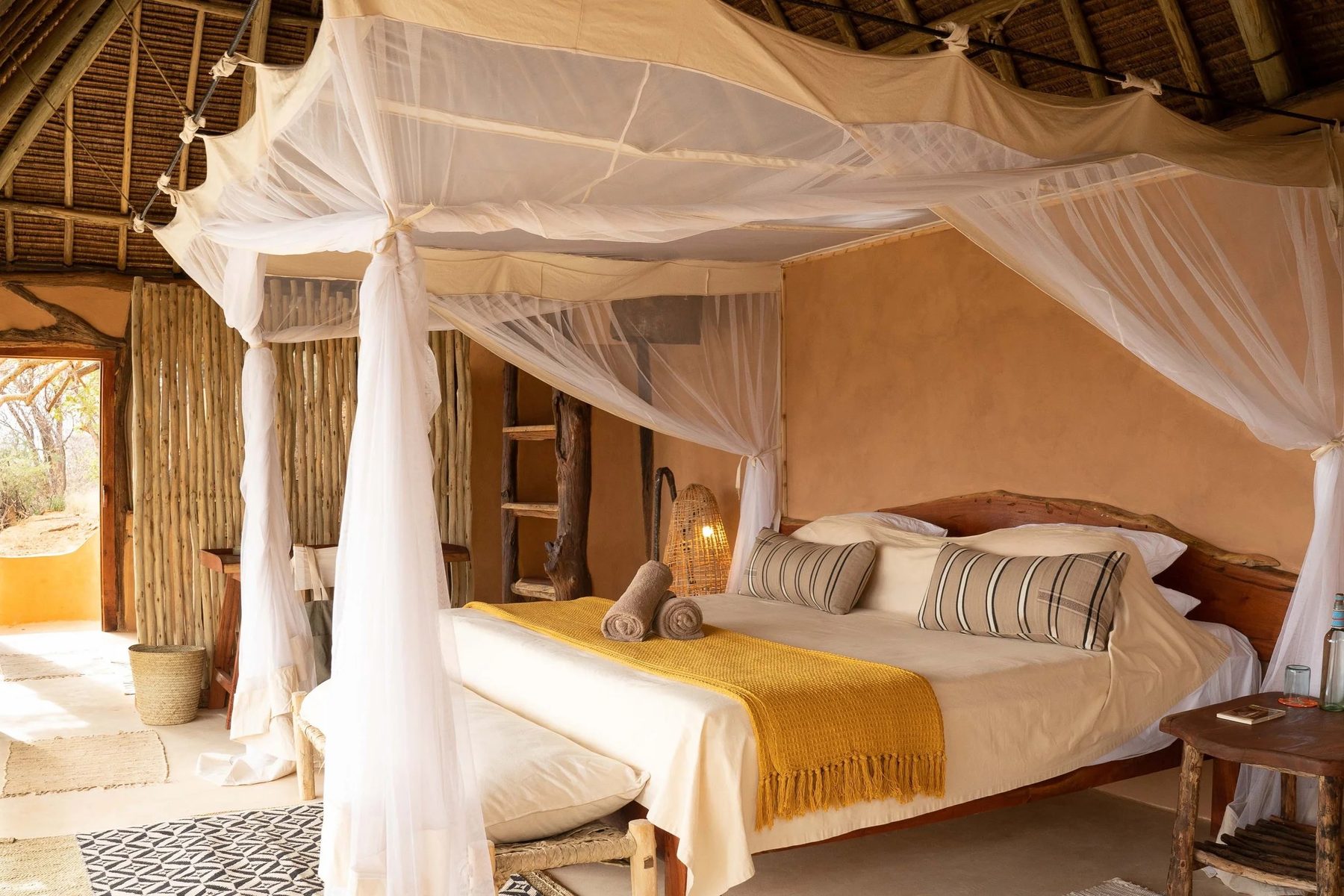 Bed under white netting with a yellow throw and striped cushions in a thatched room at Reteti House