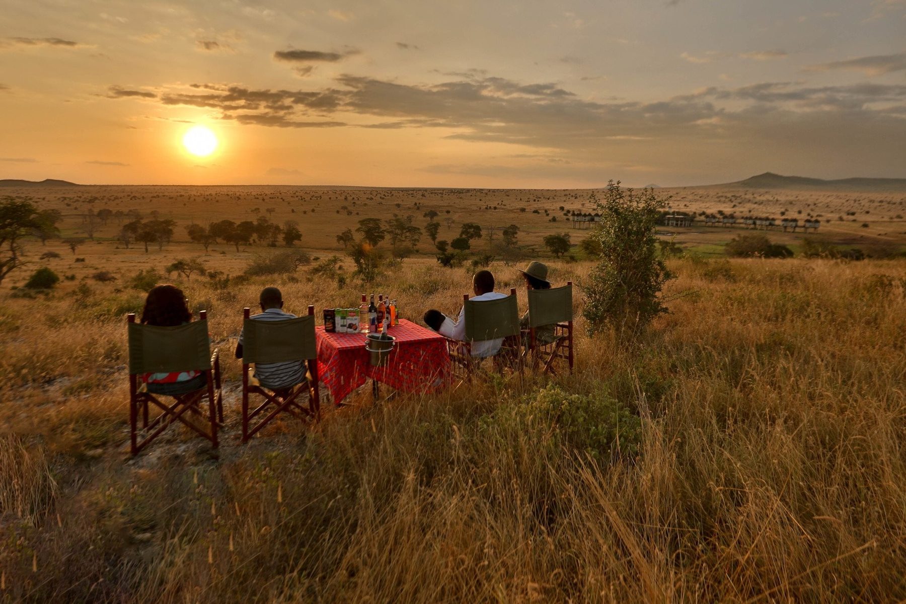 Guests in canvas chairs around a red-clothed table in the grass, watching the sun set over the plains