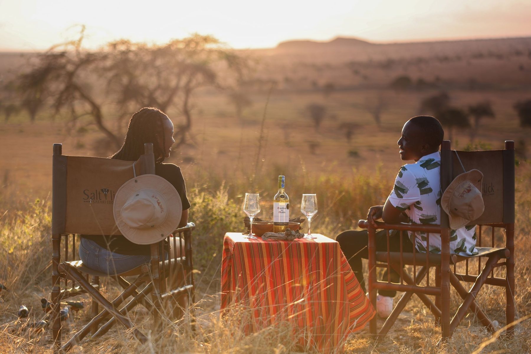 Couple in director’s chairs sharing wine at a small table in golden grass at sunset