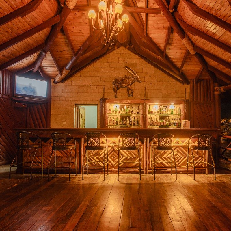 Bar at Samburu Simba Lodge with a carved wooden counter, shelves of bottles and a timber-beamed roof