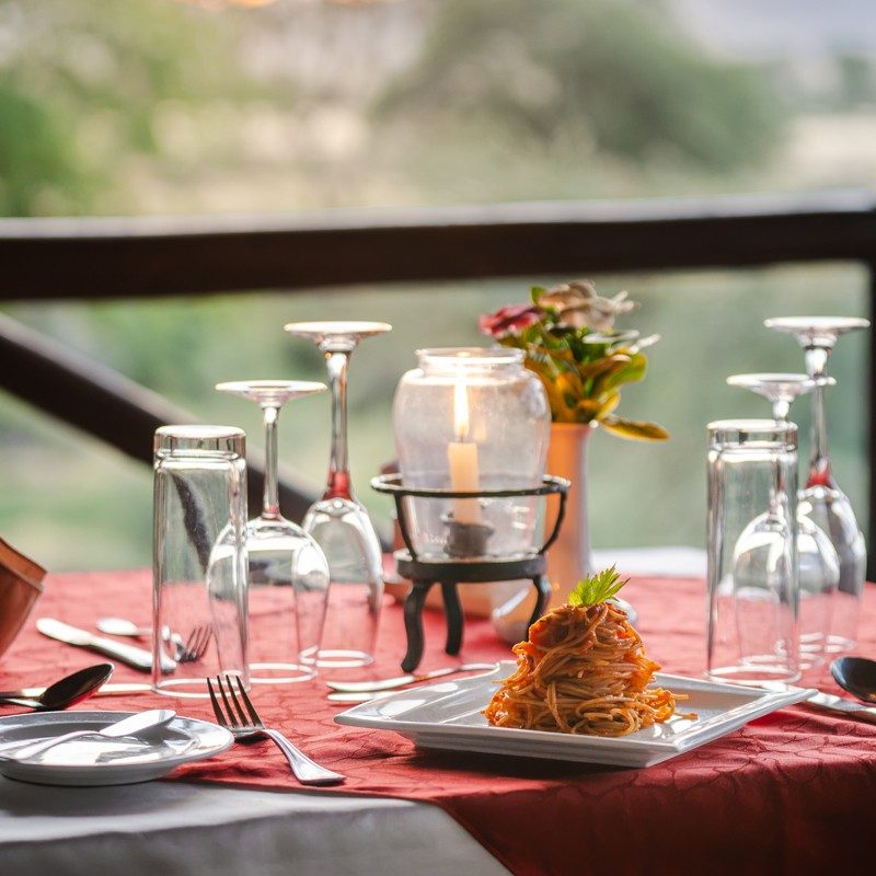 Table laid with glasses, candles and a plated meal on a red cloth at Samburu Simba Lodge, overlooking the bush