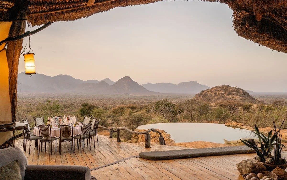Long dining table on a wooden deck beside the pool at Sarara Camp, with rocky hills and mountains beyond