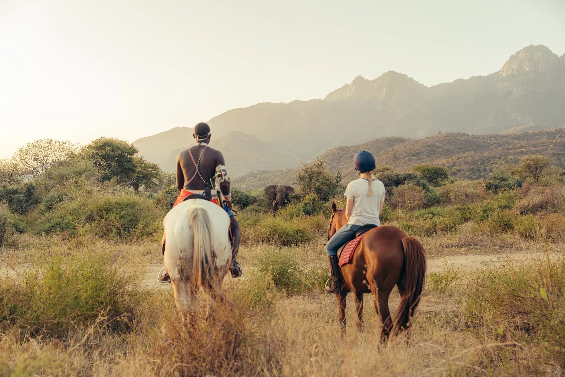 Two riders on horseback heading through dry bush towards jagged mountains near Sarara Camp