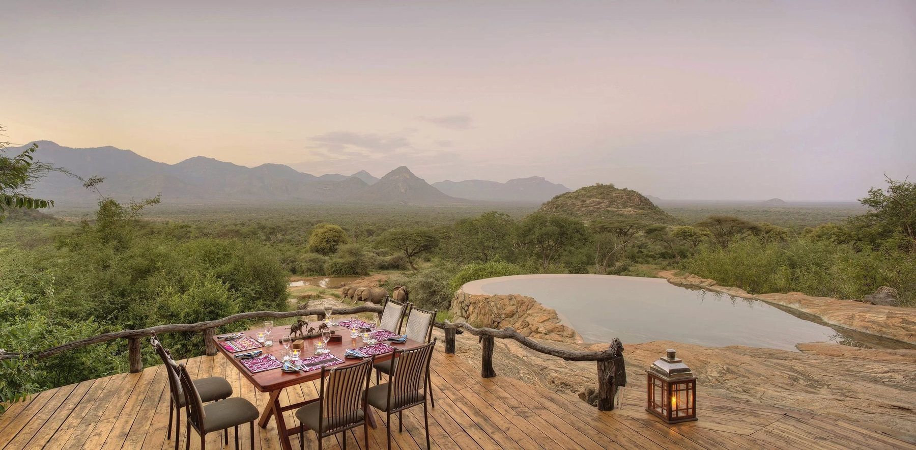 Table set on a timber deck above the rock-edged pool at Sarara Camp, overlooking a wooded valley and distant hills