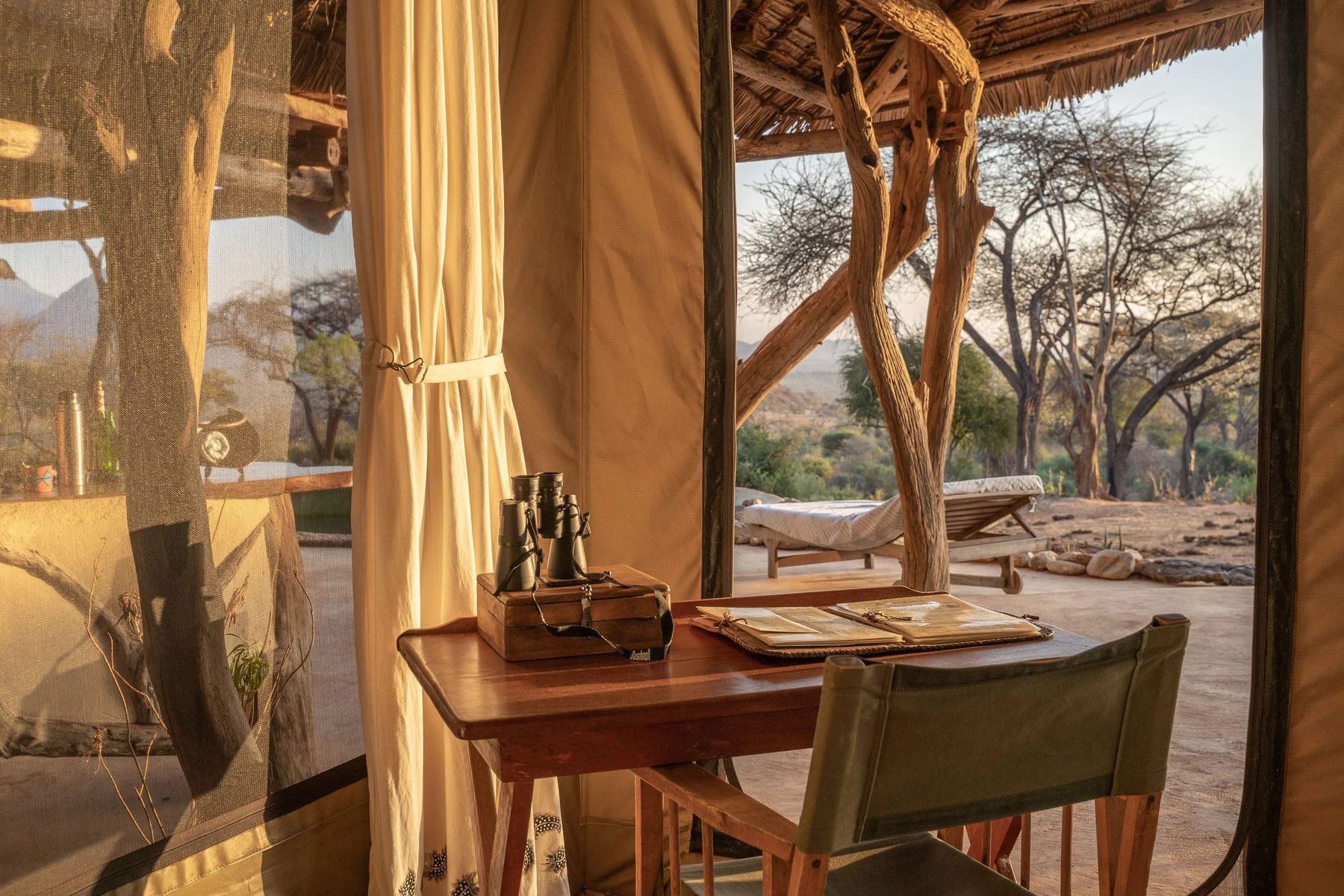 Writing desk with binoculars and a canvas chair beside the open tent flaps at Sarara Camp, looking out to acacia trees