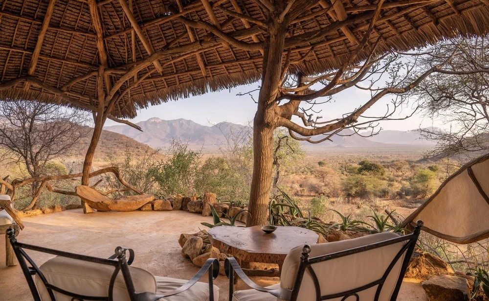 Shaded veranda with chairs under a thatched roof and a large tree at Sarara Camp, looking out to hills